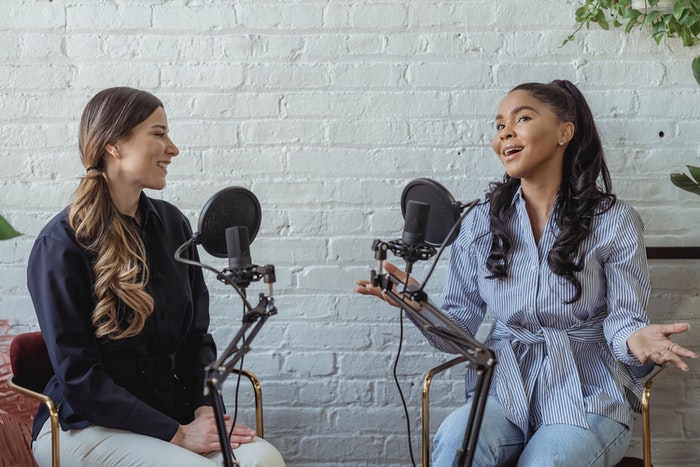 Chicas con micrófonos creando podcast Podcast: Qué es y cómo puedes crear uno para tu marca, ¿Cuántos tipos de podcasta existen?